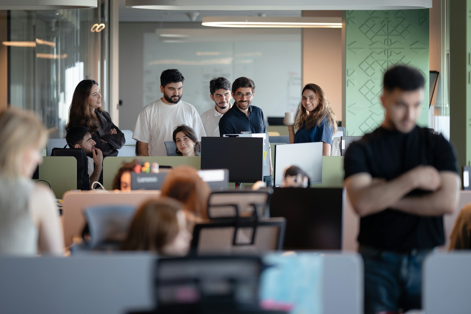 A smiling team gathered around a workstation in a busy open-plan office